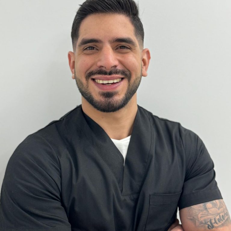 Profesional de la salud sonriente con uniforme médico, de pie frente a una pared blanca.